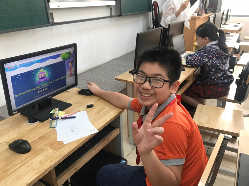A child sitting at a desk with a computer and other people in the background

Description automatically generated with low confidence